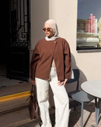 Woman in a brown jacket and white hijab standing outdoors near a building.